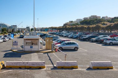 Girit, Yunanistan - 7 Nisan 2025: Girit 'te bir liman yakınında içi araba dolu hareketli bir otopark. Arka planda bir yolcu gemisi, ağaçlar ve yakınlardaki kentsel binalar yer alıyor..