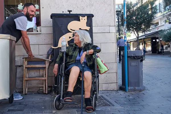 İngilizce: Girit, Yunanistan - 7 Nisan 2025: Tekerlekli sandalyede yaşlı bir insana yardım eden bir garsonun yer aldığı şehir sahnesi. Yoldan geçenlerle ve dışarıda yemek yiyenlerle canlı bir sokakta yakalanan bir nezaket anı..