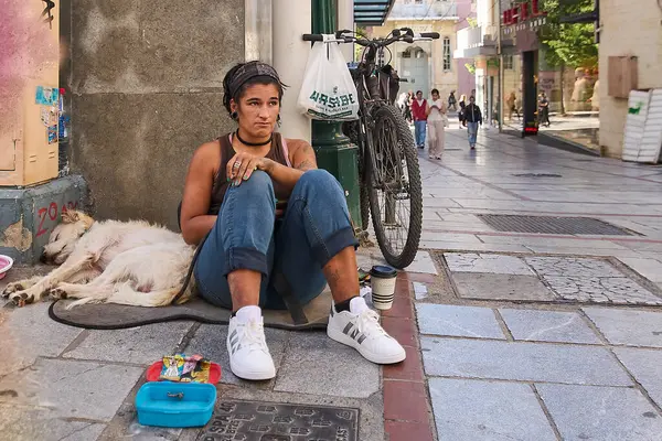 Girit. Yunanistan - 07 Nisan 2025: Sadık beyaz bir köpek yakınlarda dinlenirken sokakta oturan evsiz bir birey. Çevresindeki nesneler arasında bir bisiklet, küçük bir kap ve kahve fincanı yer alıyor.