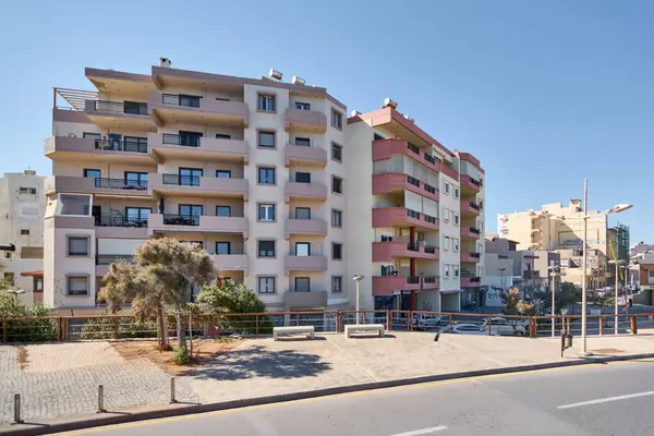 Girit. Yunanistan - 11 Nisan 2025: Girit 'te modern apartmanlar, balkonlar ve park halindeki arabaların yer aldığı güneşli bir şehir sokağı. Temiz çevre ve çağdaş mimari günlük şehir yaşamını vurgular.