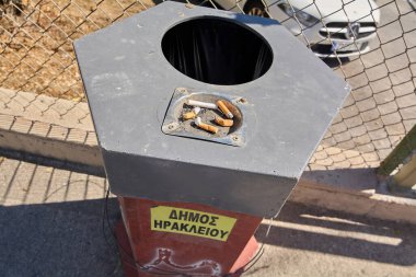 Girit. Yunanistan - 23 Nisan 2025: Heraklion 'da sokak kül tablası ve sigara öğütücü için metal bir plaka. Sarı bir belediye levhası görülebiliyor, arka planda park halindeki araçların olduğu tel örgülerin yanına yerleştirilmiş..