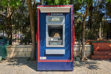 Girit. Yunanistan - 23 Nisan 2025: Eurobank ATM 'si yakındaki ağaçlı ve çitli yaya geçidine kuruldu. Mavi ve kırmızı makine kullanıcılara uygun bir dış mekanda para çekme olanağı sağlıyor.