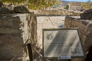 Knossos 'ta bilgilendirme plaketi, geçit töreni koridoru ve sarayın batı verandası. Tabelada tarihi gerçekler, site şemaları ve arkeolojik referanslar yer alıyor.