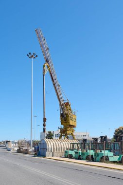 Girit. Yunanistan - 05 Mayıs 2025: Demiryolu üzerinde devasa sarı bir vinç ve düzgün düzenlenmiş forkliftlerin yer aldığı çitli bir alanda endüstriyel ekipman. Uzun ışık direkleri çalışma alanını aydınlatıyor ve gökyüzü açık.