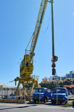 Girit. Yunanistan - 05 Mayıs 2025: Altında çok sayıda yeşil forkliftin park edildiği bir sanayi bölgesinde sarı bir vinç duruyor. Çitle çevrili alan büyük ölçekli malzeme işleminin bir parçası.