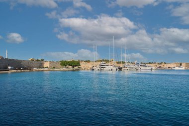 Girit. Yunanistan 15 Mayıs 2025: tarihi mimariyle çevrili ve dağınık bulutlu canlı mavi bir gökyüzü bulunan birkaç yatı demirlemiş sakin Rhodes limanı suları.