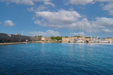 Yunanistan 15 Mayıs 2025: Lüks yatlar, berrak mavi sular ve parçalı bulutlu gökyüzü altında antik tahkimatların yer aldığı Rhodes Marina manzarası seyahat ve turizm için mükemmel.