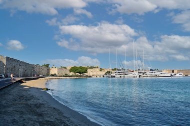 Kavisli kumlu bir sahil manzarası, rıhtımlı yatlar ve güneşli bir gökyüzünün altında engebeli tarihi taş duvarlar. Seyahat, tarih veya Akdeniz turizm temaları için ideal. Canlı renkler ve yansıtıcı su görsel cazibeyi artırır.