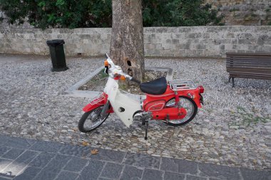 Rodas.Yunanistan - Haziran 01, 2025: Güzelce korunmuş bir retro scooter, tarihi mimari ve yemyeşil arkaplan karşısında ikonik tasarımını sergileyerek, dokulu bir kaldırım taşı caddesinde dinleniyor..