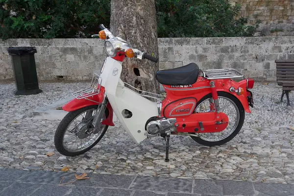 Rodas.Yunanistan - Haziran 01, 2025: Arka planda antik taş bir duvar bulunan geleneksel bir kaldırım taşı sokağındaki bir ağacın yanına çarpan kırmızı-beyaz bir Honda Super Cub scooter park edilmiştir..