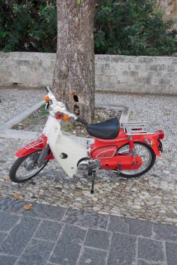 Rodas.Yunanistan - Haziran 01, 2025: klasik kırmızı ve beyaz moped, tarihi kent dekoruna biraz nostaljik cazibe katıyor, eski taş duvarlar ve gölgeli asfalt ile tamamlanmış.
