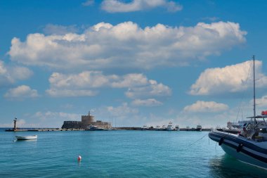 Rodas.Yunanistan - Haziran 04, 2025: Rodos 'taki Picturesque Mandraki Limanı, tarihi Saint Nicholas Kalesi, lüks yatlar ve parlak gökyüzü altındaki berrak gök mavisi sular.