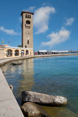 Rodas.Yunanistan - Haziran 04, 2025: Mandraki Limanı 'ndan Rodos' un heybetli saat kulesi, sakin suları yansıtan önemli bir dönüm noktası..