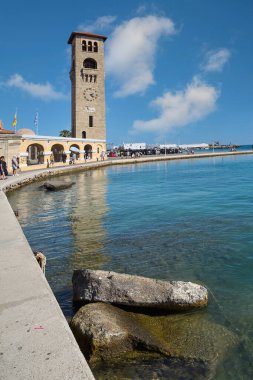 Rodas.Yunanistan - Haziran 04, 2025: Rodos 'un ikonik saat kulesi Mandraki Limanı' nın yanında duruyor, dingin suları parlak mavi gökyüzünü ve beyaz bulutları yansıtıyor..