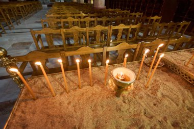 Kum dolu bir stantta mumlar yakmak, Annunciation Kilisesi 'nde sadakati ve duayı sembolize ediyor, arka planda ahşap banklar var..