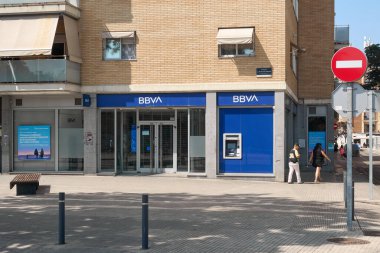 Viladecanlar. Barcelona - 16 Temmuz 2025: Bir BBVA bankasının önündeki hareketli şehir kaldırımının günlük yaşantısını ve finansal erişilebilirliğini gösteren gündüz manzarası.