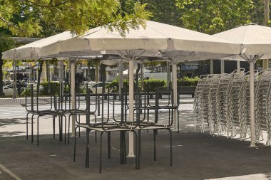 Viladecans.Barcelona - 19 Temmuz 2025: Masaları ve sandalyeleri büyük şemsiyelerin altına istiflenmiş kapalı kafe terası. Müşterilerin acele etmesinden önce ya da sonra kentsel alanın sakinliği, sessiz bir momet.