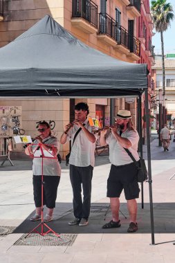 Viladecanlar, Barcelona - 25 Temmuz 2025: Çadırın altında rüzgar enstrümanları çalan üç müzisyen. Canlı kentsel etkinlik sahnesi, eğlence ve eğlence için ideal..