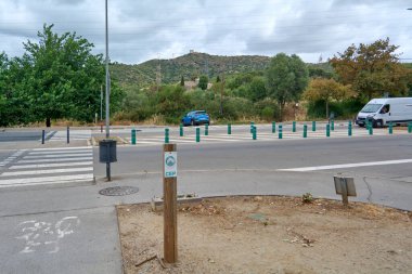 Viladecans.Barcelona - 28 Temmuz 2025: Yeşil cüsseli ve yaya geçitli bir yol kavşağı, görülebilir binaları olan doğal bir dağ manzarasına doğru gidiyor..