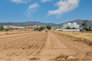 Viladecanlar. Barselona - 14 Ağustos 2025: Arka planda paralel çizgiler, dağlar ve kentsel gelişimi sembolize eden modern binaların bulunduğu bir toprak arazinin görüntüsü.