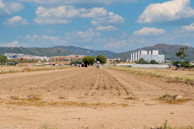 Viladecanlar. Barselona - 14 Ağustos 2025: Arka planda paralel çizgiler, dağlar ve kentsel gelişimi sembolize eden modern binaların bulunduğu bir toprak arazinin görüntüsü.