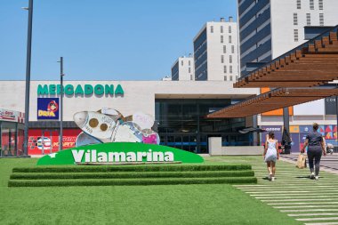 Viladecanlar. Barselona - 29 Ağustos 2025: Mercadona süpermarketinin ve açık hava meydanında yürüyen insanların yer aldığı Vilamarina alışveriş merkezinin ana girişi.
