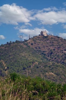 Sant Ramon 'un Hermitage' ı kuru bir tepenin üstünde gösteriliyor. Bulutların ve elektrik hatlarının varlığı manzaraya biraz dinamizm katıyor..