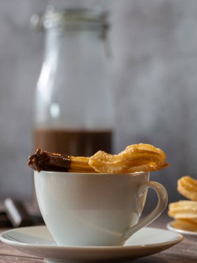 Çikolata kaplı churro 'nun üstüne beyaz bir fincan sıcak çikolata. Arka planda çikolata olan cam şişe. 