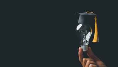 academy and success graduate education concept. businessman hand holding bright, electric light bulb with degree cap on black background. business education, knowledge, learning idea with copy space.