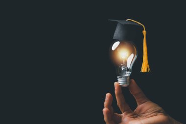 academy and success graduate education concept. businessman hand holding bright, electric light bulb with degree cap on black background. business education, knowledge, learning idea with copy space.