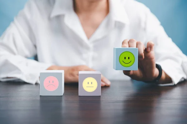 Woman hand holding happy face smile face icon on cube blue object. Customer experience and service with satisfaction concept. positive thinking, mental health assessment, world mental health day.