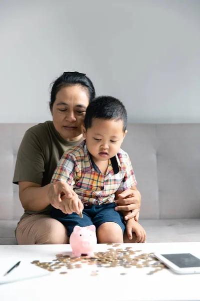 Happy Asian kid and mom saving money together, putting cash into piggy bank. Mother playing with child on heating floor at home, teaching little son to invest money, planning future.