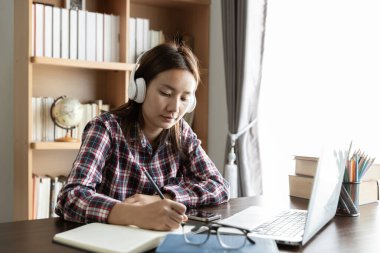 Asyalı kız öğrenci internetten Skype öğretmeniyle kablosuz kulaklık çalışması yapıyor, mutlu genç kadın dil öğreniyor. Ders veriyor, web sitelerini izliyor, not yazıyor, dizüstü bilgisayara bakıyor, evde oturuyor, uzaktan eğitim görüyor.