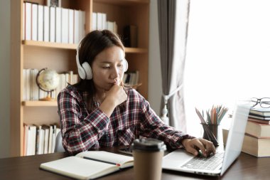 Asyalı kız öğrenci internetten Skype öğretmeniyle kablosuz kulaklık çalışması yapıyor, mutlu genç kadın dil öğreniyor. Ders veriyor, web sitelerini izliyor, not yazıyor, dizüstü bilgisayara bakıyor, evde oturuyor, uzaktan eğitim görüyor.