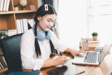 Asyalı kız öğrenci kulaklık takıyor internetten öğreniyor Webinar dersini izliyor bilgisayarlara bakıyor uzaktan ders alıyor notlar alıyor ya da evde sanal konferans öğretmeniyle görüşüyor..