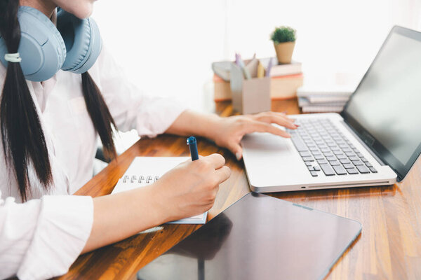 Asian girl student wear headphones learning online watching webinar class looking at laptop computer elearning remote lesson making notes or video calling virtual conference meeting teacher at home.
