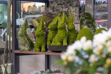 Raftaki eğlenceli, sevimli, yosunlu, yeşil Paskalya tavşanları ailesi. Yosundan yapılmış büyük yeşil Paskalya yumurtaları. Önplanda beyaz bulanık çiçekler.