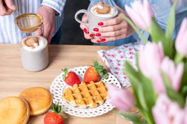 Kırmızı tırnaklı açık mavi elbiseli bir erkek ve bir kadının elleri. Gri bardakta tarçın ve sütlü kahve reklamları yapan bir adam. Çıtır waffle, beyaz tabakta kırmızı çilek ve donut..