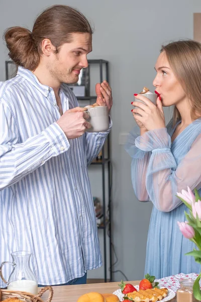 Güzel, gülümseyen, kalın saçlı bir adam ve kırmızı dudaklı ve tırnaklı güzel genç bir kadın, ikisi de açık mavi elbiseli, kahve içiyor ve birbirlerine bakıyorlar..