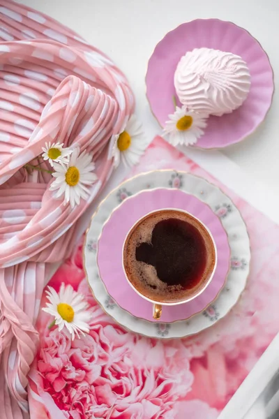 Çiçekli pembe bir peçetenin üzerinde nefis kırılgan bir kahve fincanında espresso kahvenin üst görüntüsü ve pembe bir fincan tabağında şişkin havadar marshmallow, şık bir polka nokta atkısı ve masada güzel, güneşli papatya çiçekleri.