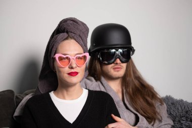 Kırmızı dudaklı genç bir kadın pembe güneş gözlüğü takıyor, uzun saçlı genç bir adam motosiklet gözlüğü takıyor, kafasında koyu gri bir kask var..