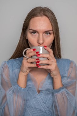 Açık mavi elbiseli güzel bir kadın, muhteşem uzun saçları, kırmızı dudakları ve tırnakları var, parmağında bir yüzük, kahvenin tadını çıkarıyor ve kameraya bakıyor..