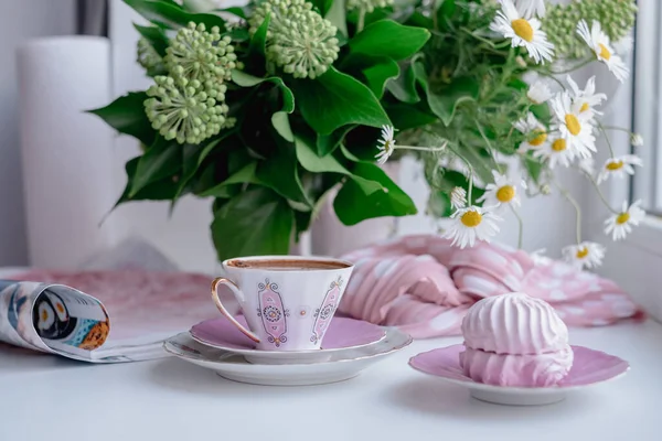 A gorgeous fragile white pink coffee cup and puffy airy pink marshmallow in a saucer on a white table. Stylish pink dot scarf, magazine and beautiful sunny daisies in white vase on background.