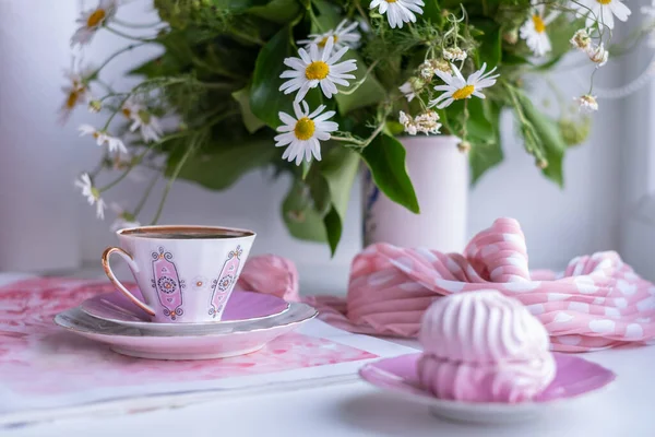 A gorgeous fragile white-pink coffee cup on a large soft pink napkin and a puffy airy pink marshmallow in a saucer on a white table. Stylish polka dot scarf and beautiful sunny daisies in white vase.