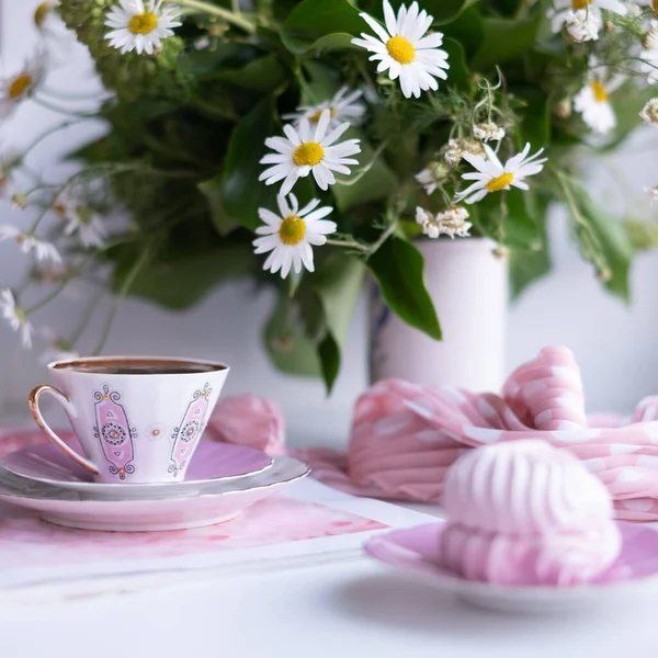 A gorgeous fragile white-pink coffee cup on a large soft pink napkin and a puffy airy pink marshmallow in a saucer on a white table. Stylish polka dot scarf and beautiful sunny daisies in white vase.