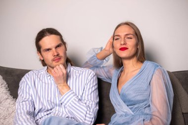 Çizgili tişörtlü düşünceli bir adam ve uzun saçlı, kırmızı dudaklı, güzel, gülümseyen bir kadın bir koltukta oturuyor ve kameraya bakıyor..