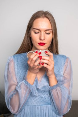 Açık mavi elbiseli, güzel, bulanık, genç bir kadın. Uzun saçları, kırmızı dudakları ve tırnakları var. Bir fincana bakıyor ve kahvenin kokusunun tadını çıkarıyor. Yüksek kalite fotoğraf