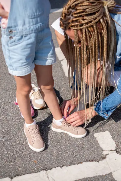 A mother with African braids pulls shoes for a girl in the parking lot. Copy space. High quality photo