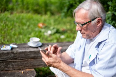 Gri saçlı ve açık mavi tişörtlü yaşlı bir adam, modern teknoloji ve son sınıf hayatının kesişimini sembolize ederek, doğadaki akıllı telefonuna dikkatle odaklanmıştır. Yüksek kalite fotoğraf