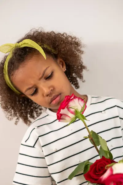 A contemplative mixed race young girl with curly hair and yellow headband smells a red rose. innocence and reflection, ideal for mothers day, first day of school, 1st September.. High quality photo